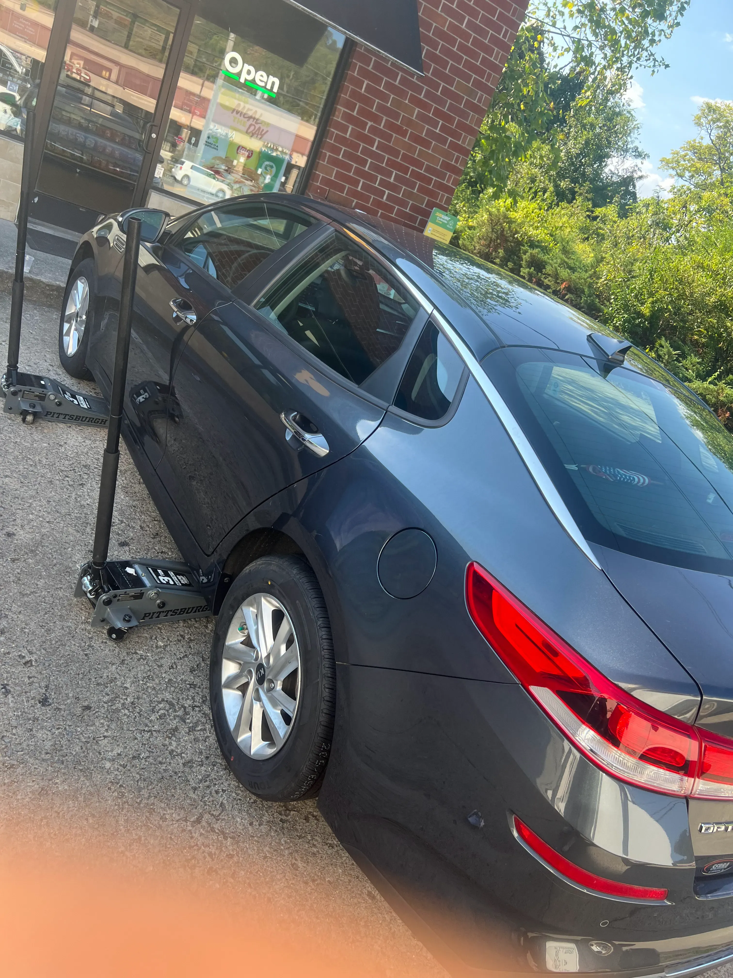 Nashville Flat Tire Service technician performing a mobile flat tire repair on a gray Kia Optima sedan with the rear wheel lifted on a hydraulic jack at a Greater Nashville apartment complex.